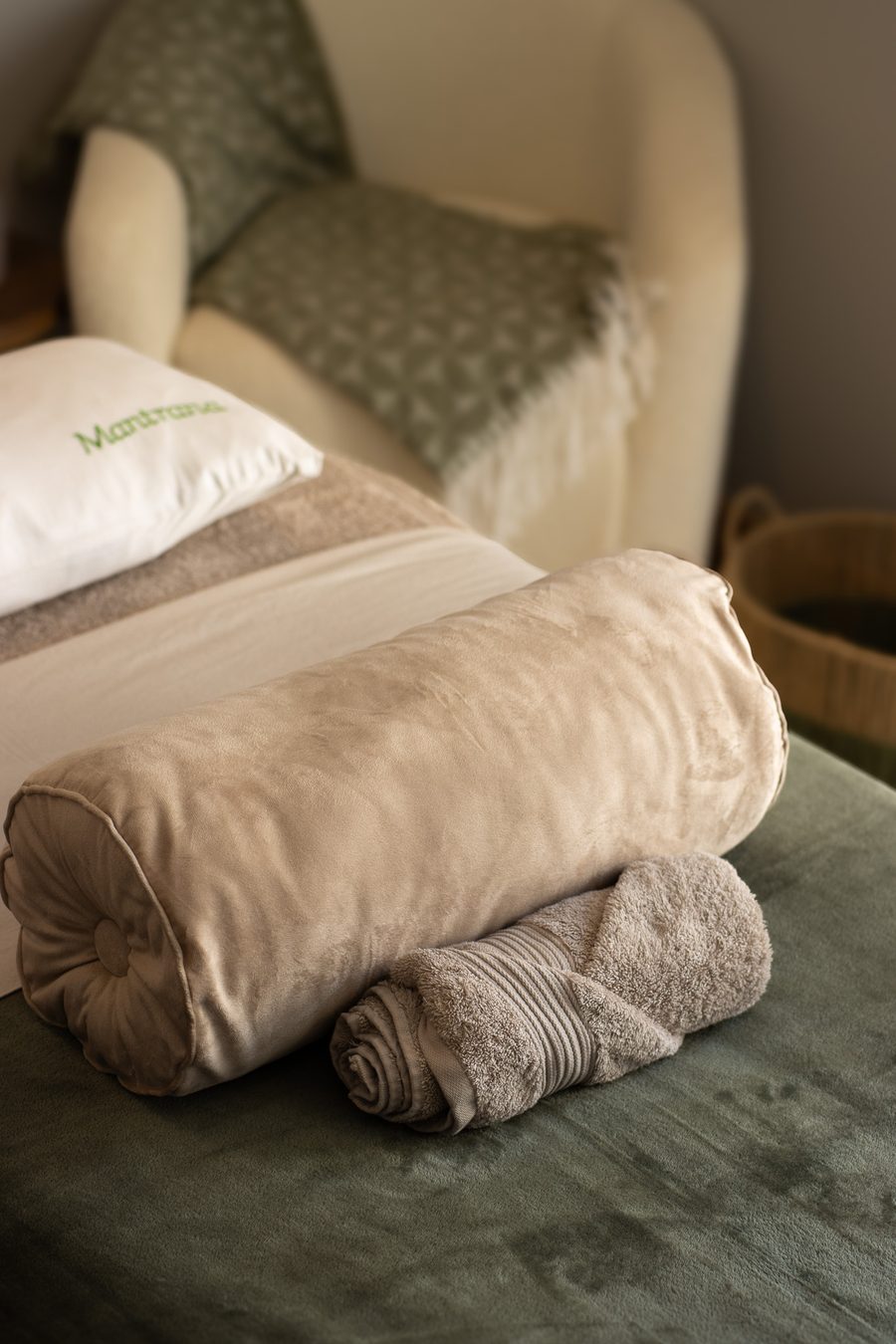 A velvet bolster and rolled towel on the treatment bed, with a cream armchair and sage throw behind