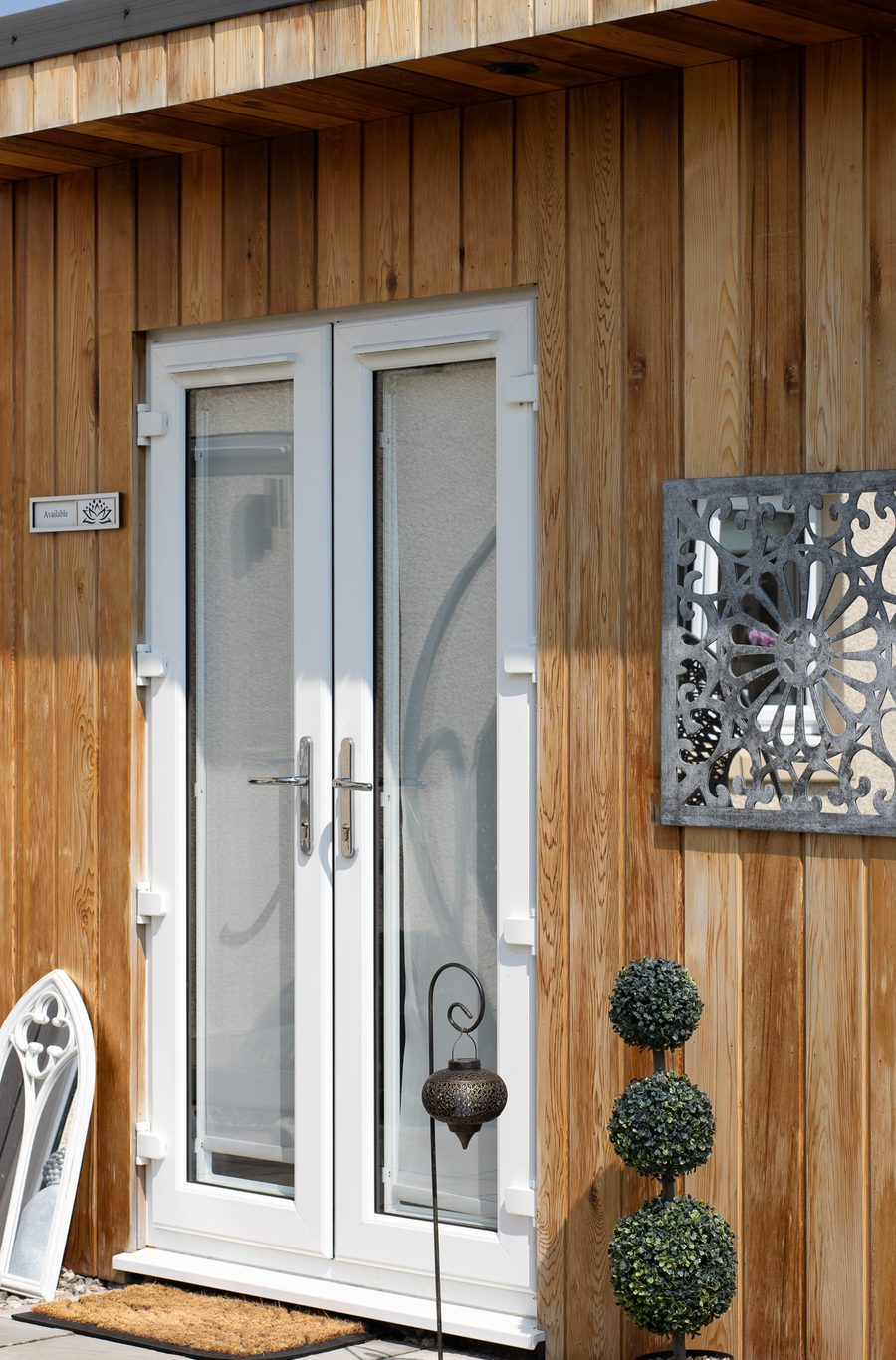 The timber-clad garden cabin with white glazed double doors, a lantern and topiary by the step