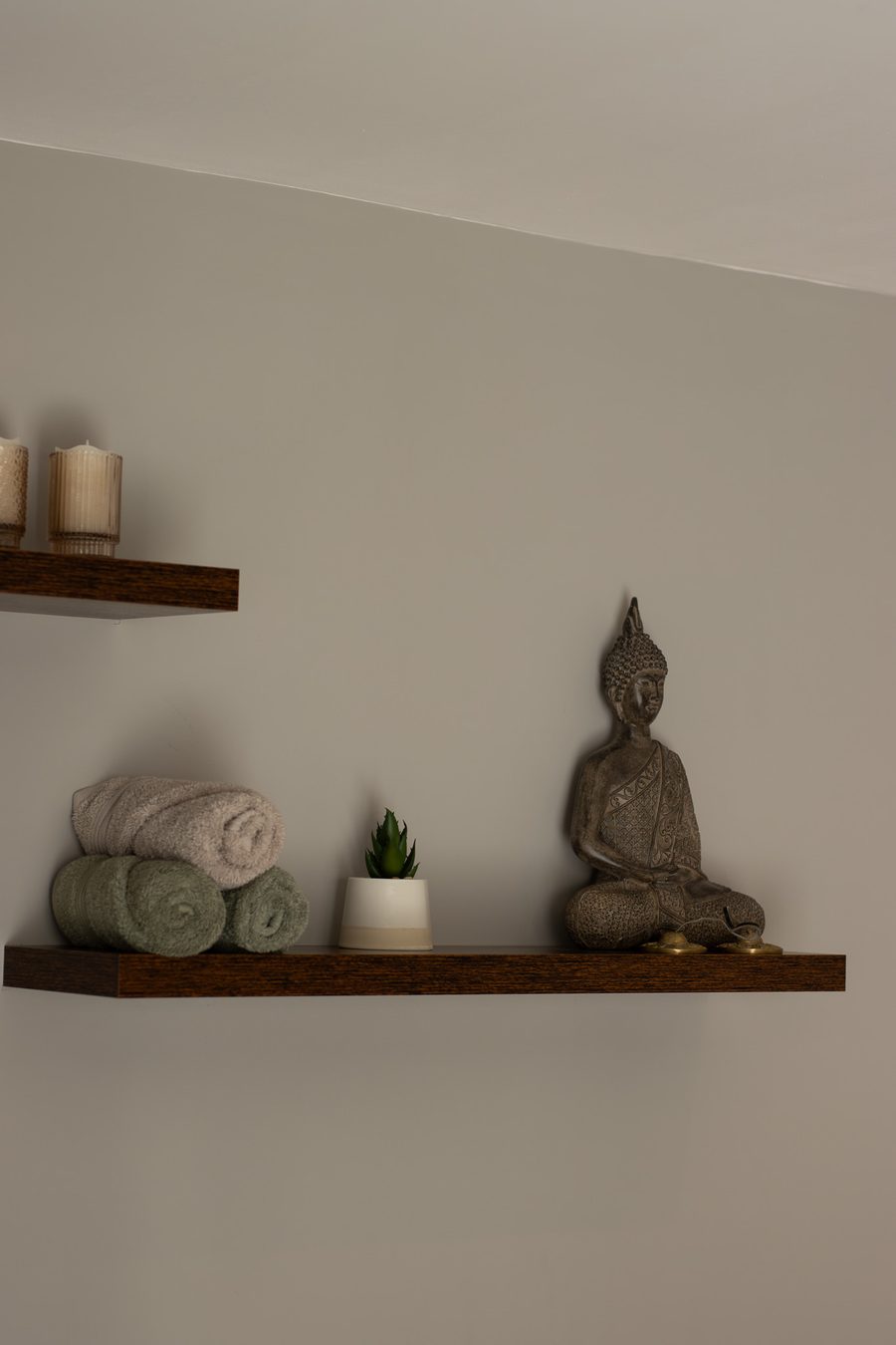 A walnut floating shelf with rolled towels, a small plant and a seated Buddha